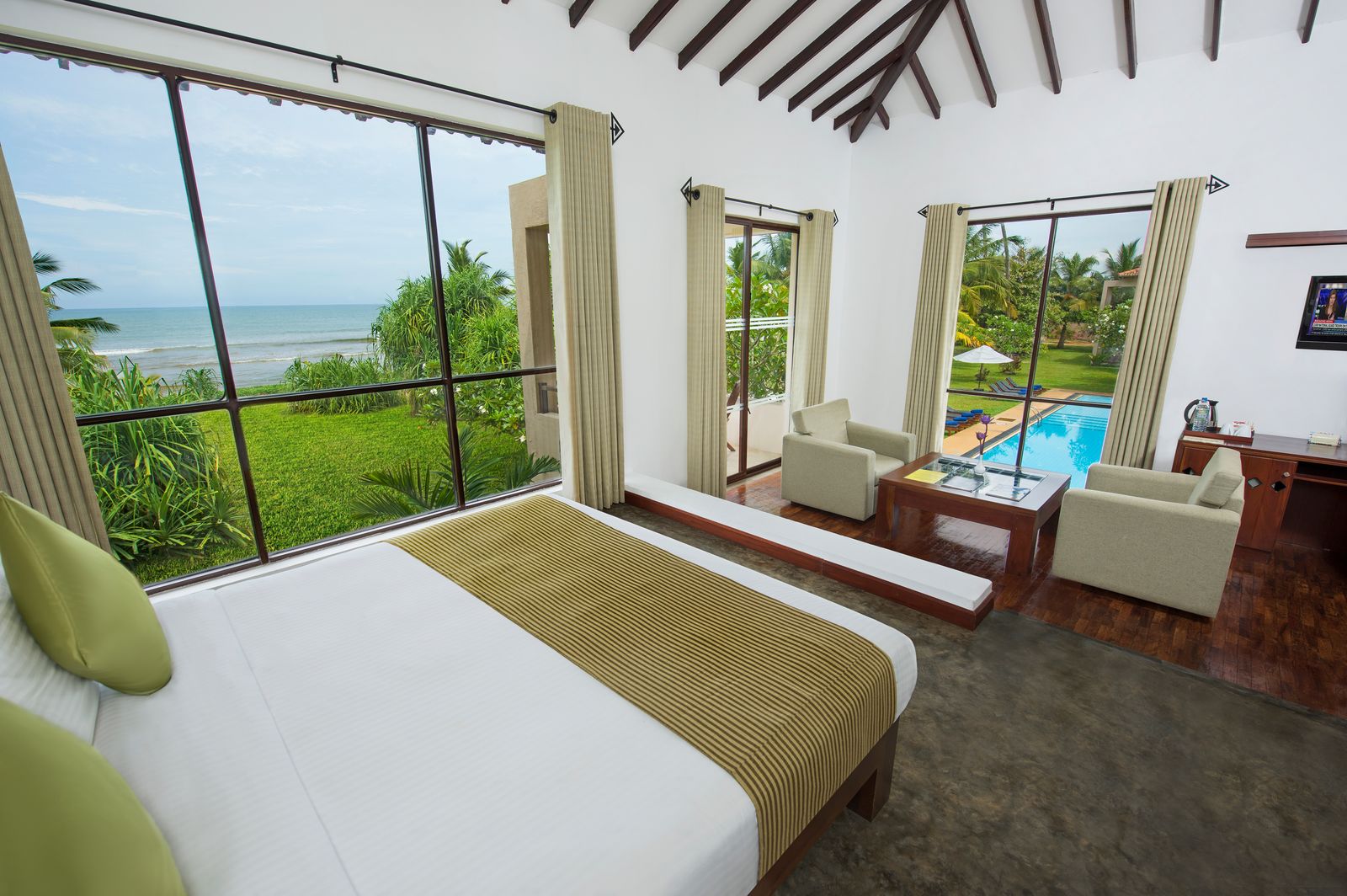 A Superior Sea View room under a sloping beamed ceiling: a double bed beside a large window looking over the garden to the sea, 2 armchairs, and a glass door onto a pool